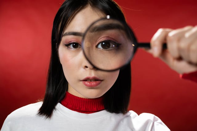 A woman in a red turtleneck sweater holding a magnifying glass close to her eye.