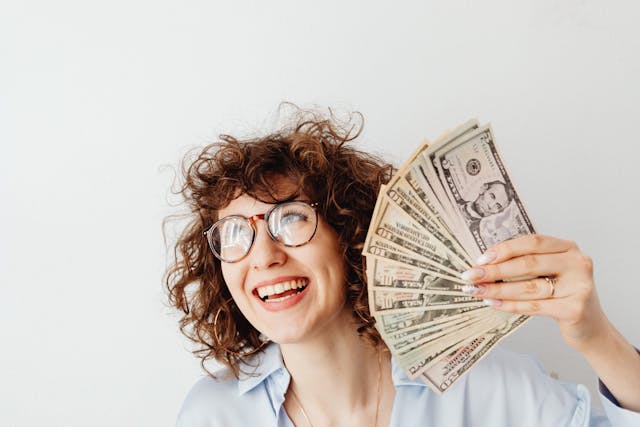 A woman with curly hair and glasses smiling while holding up a fan of cash.