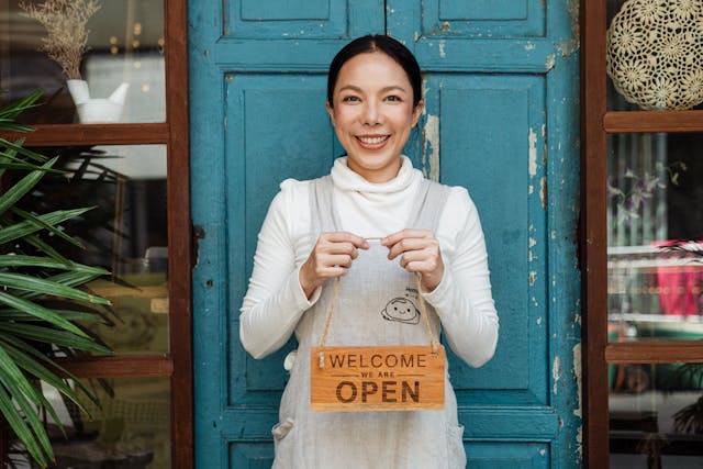 A café owner in a gray apron smiling and holding a welcome sign outside her business.