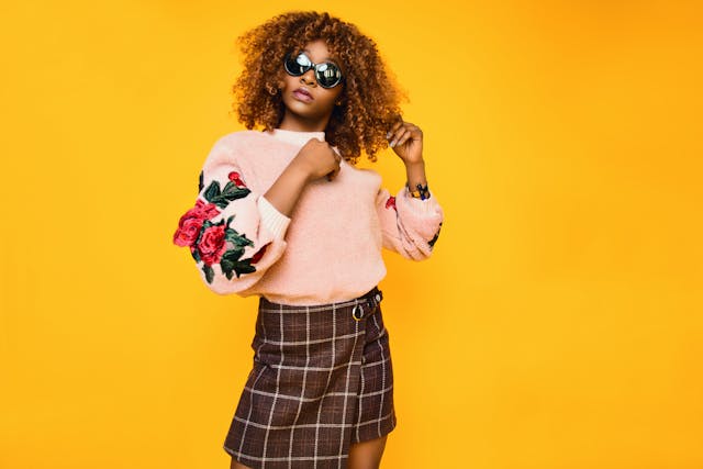 A woman with sunglasses, a pink sweater, and a brown plaid skirt posing against a yellow background.