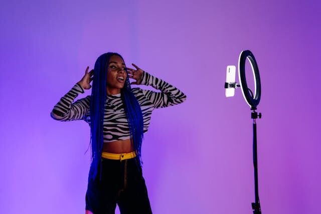 A woman with blue braids and trendy clothes posing in front of a phone camera and ring light.