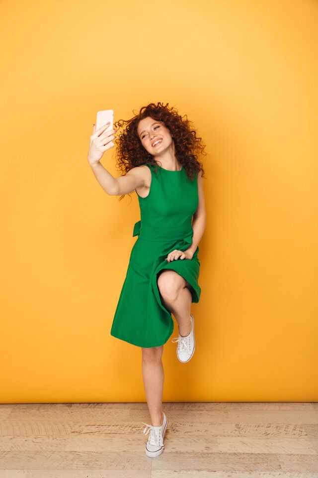 A lively female influencer with curly hair and a green dress smiling and posing while taking a selfie.