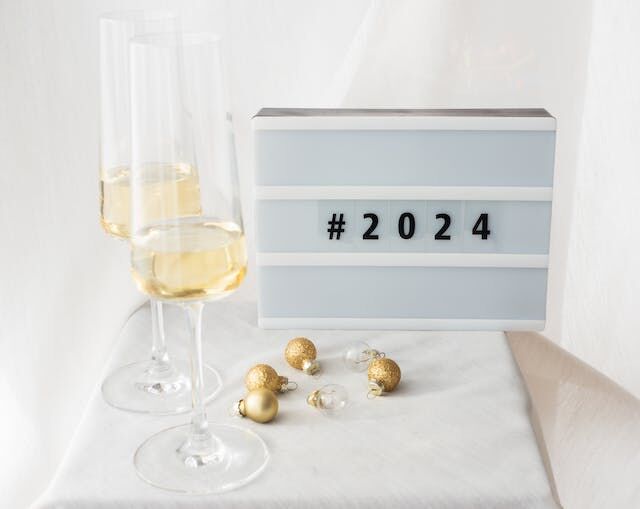 Een bord met de tekst #2024 naast twee glazen en kerstversieringen op een tafel.