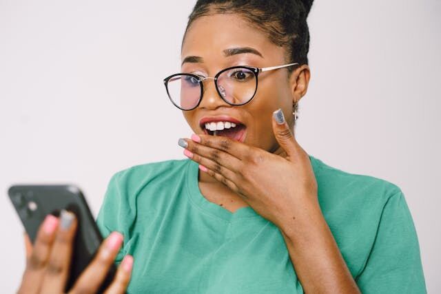 Eine Frau hält sich den Mund zu und macht ein schockiertes Gesicht, während sie etwas auf ihrem Handy sieht.